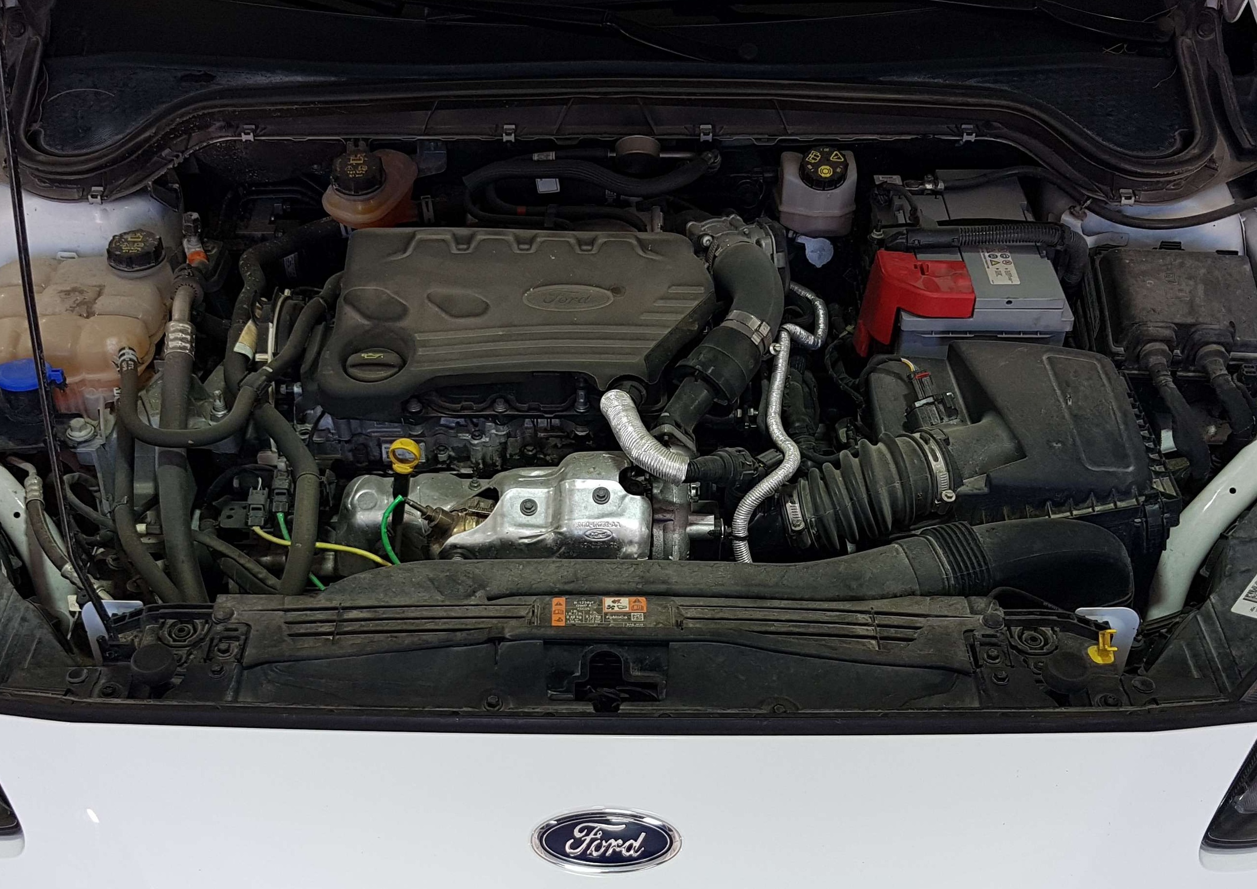 Front view of a car engine bay under the open hood, Ford engine cover visible with battery to the right and various hoses.